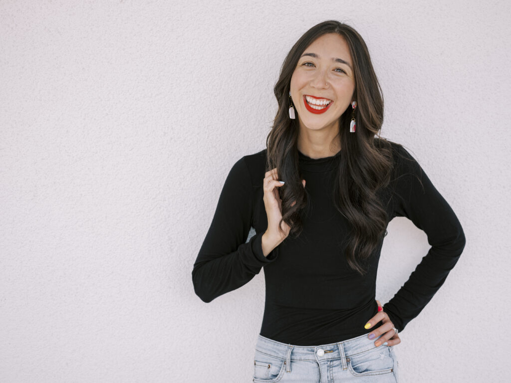 woman in a black sweater smiling at the camera with bright red lipstick in front of a white wall