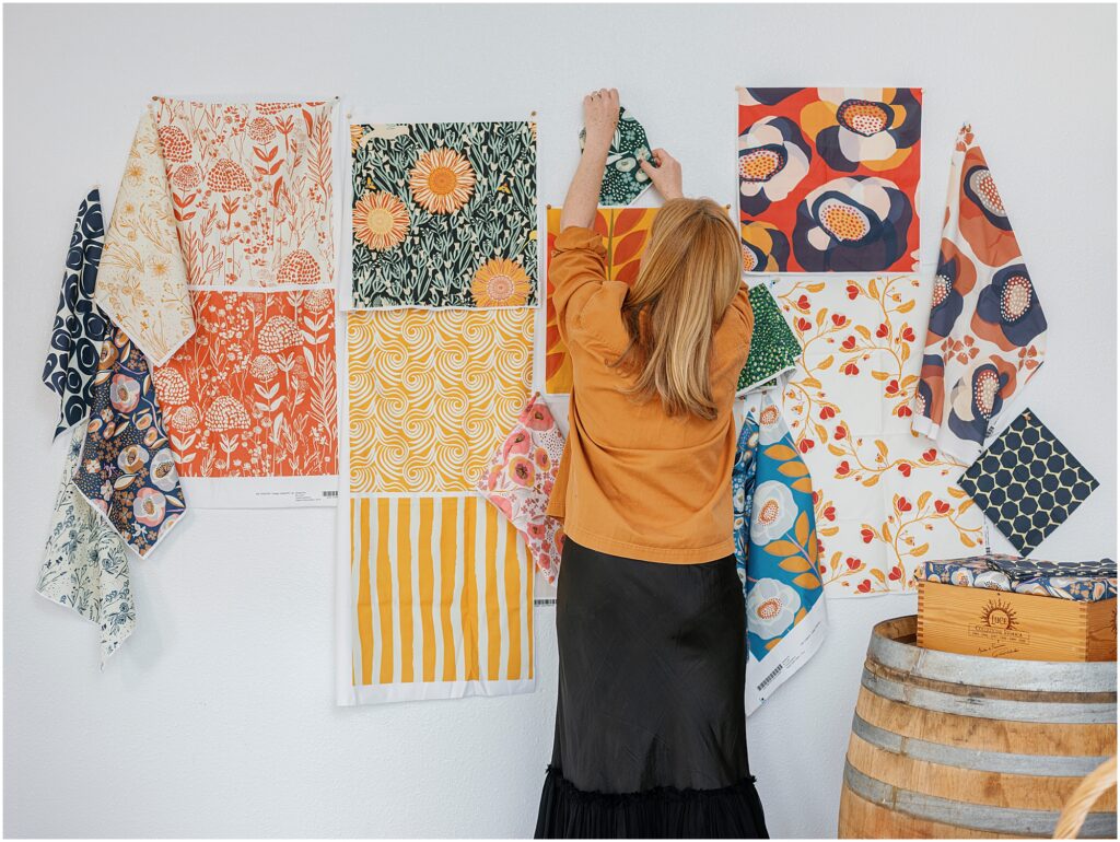 woman pinning orange and red and green fabric swatches with floral prints to a wall photographed by Krista Marie Photography, a Bay Area personal brand photographer